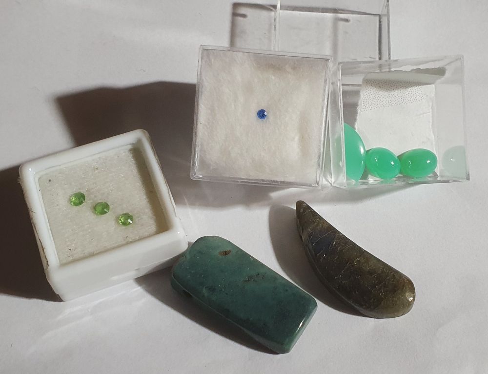 Blaue und grüne Edelsteine auf weißem Papier. Hintere Reihe: 3 geschliffene Olivine in einem Kästchen,  1 intensiv blauer Hauyn,  3 Chrysopras-Cabochons. Vorne 2 massive polierte Anhänger mit Bohrung: rechts ein Labradorit in Paisley-Form. Was der mattgrüne Stein links ist, weiß ich nicht mehr. 
