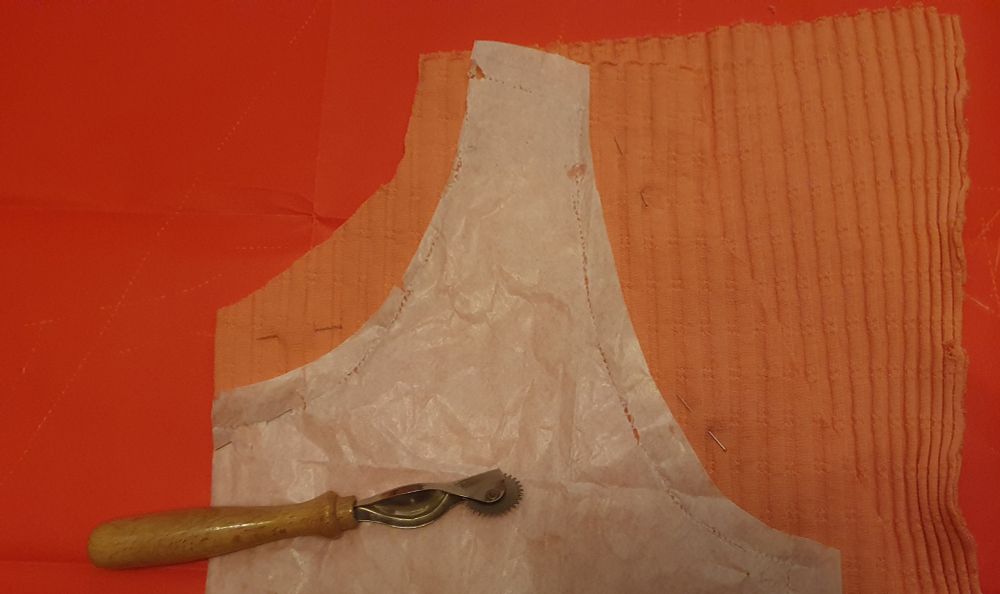 Ein Schnittmuster und ein Kopierrädchen liegen auf einem lachsrosa Jerseystoff, darunter rotes Kreidepapier .