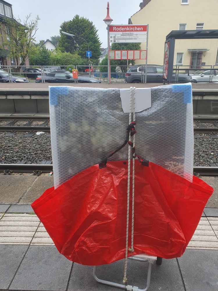 Ein Foto vom Rodenkirchener Bahnhof. Auf dem Bahnsteig steht ein Shopper, darauf eine große rote Plastiktasche mit einem eingepackten Bild drin.