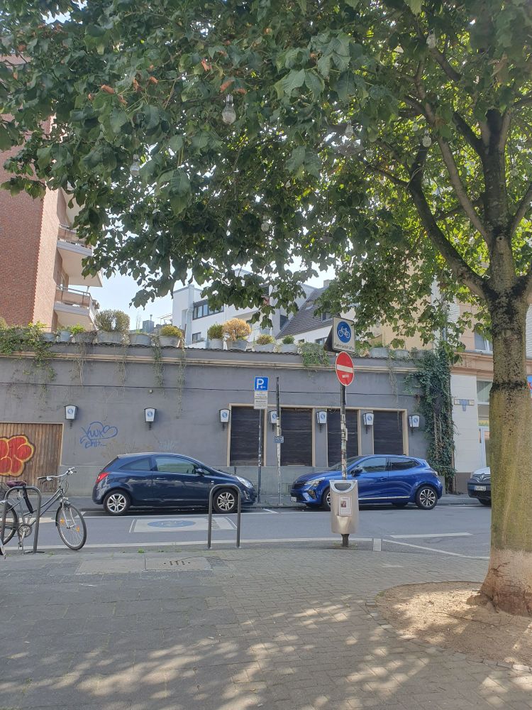 Ein schattiger Platz unter einem Baum, durch Poller und ein Straßenschild abgeriegelt. In der kleinen Straße parken 2 blaue Autos. Die Gaststätte dahinter hat einen Dachgarten im 1. Stock. Dahinter weitere Häuser, Balkone... Rückseiten von Stadhäusern.