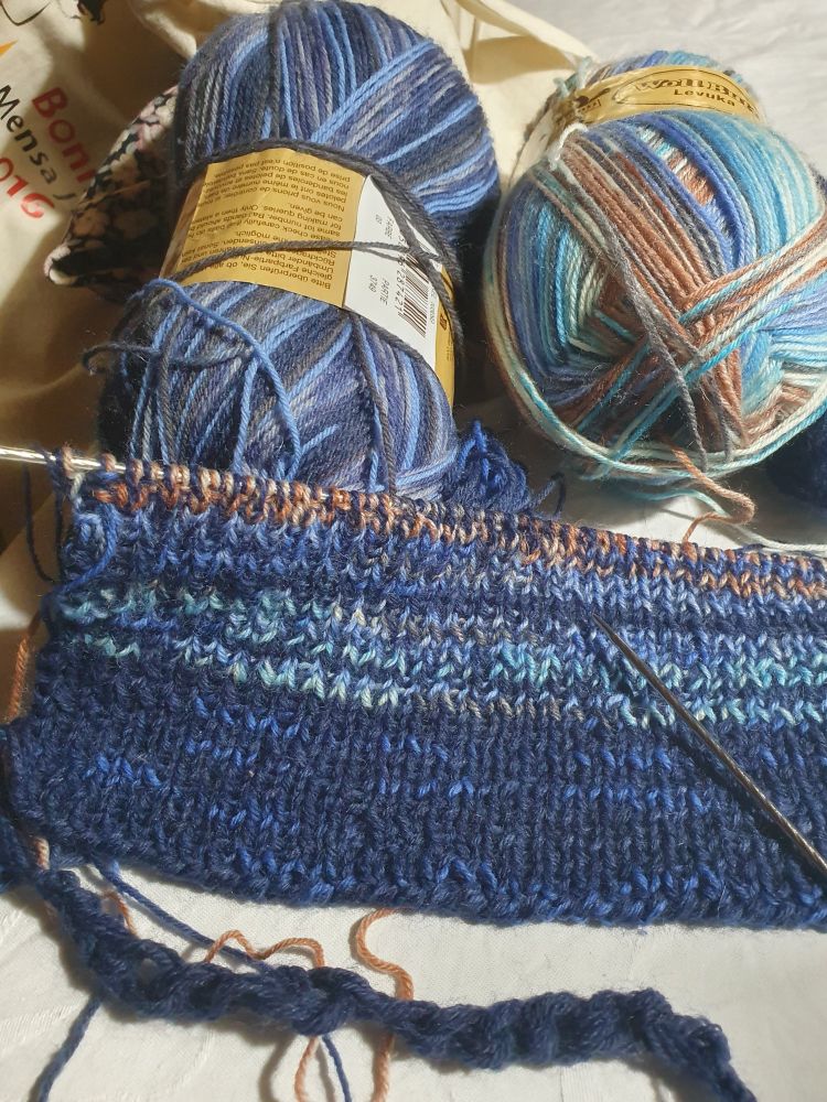 Eine angefangene Strickarbeit in Blautönen. Zwei dicke Knäuel liegen hinter dem Gestrick. 