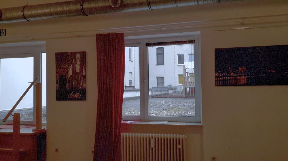 Blick aus dem 'Salon' auf den Hinterhof mit hellen Wänden und Fenstern, gekiestem Garagendach und umlaufendem Rohr  über den Fenstern. Neben den Fenstern 2 Bilder: das gelbe Fahrrad vor dem Dom und ein Bild von den Kranhäusern in der Dunkelheit, während sich Lichter im Rhein spiegeln.
