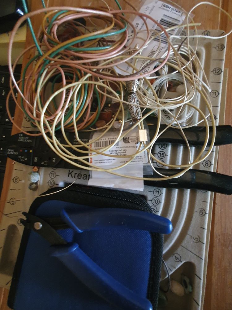 Auf einem Perlbrett liegen: Werkzeugtasche, Zange, bunte Lederschnüre, verschiedene Perlen. 
