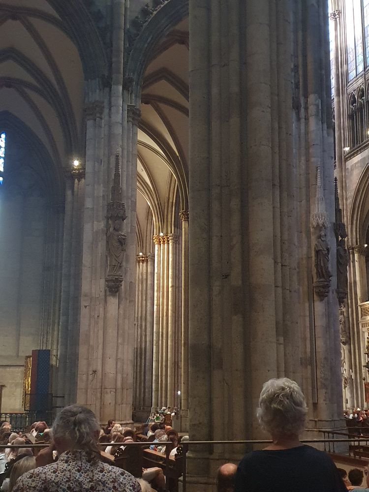 Blick ins Seitenschiff des Kölner Doms mit Steinsäulen. Einige Zuhörer von hinten am unteren Rand. 