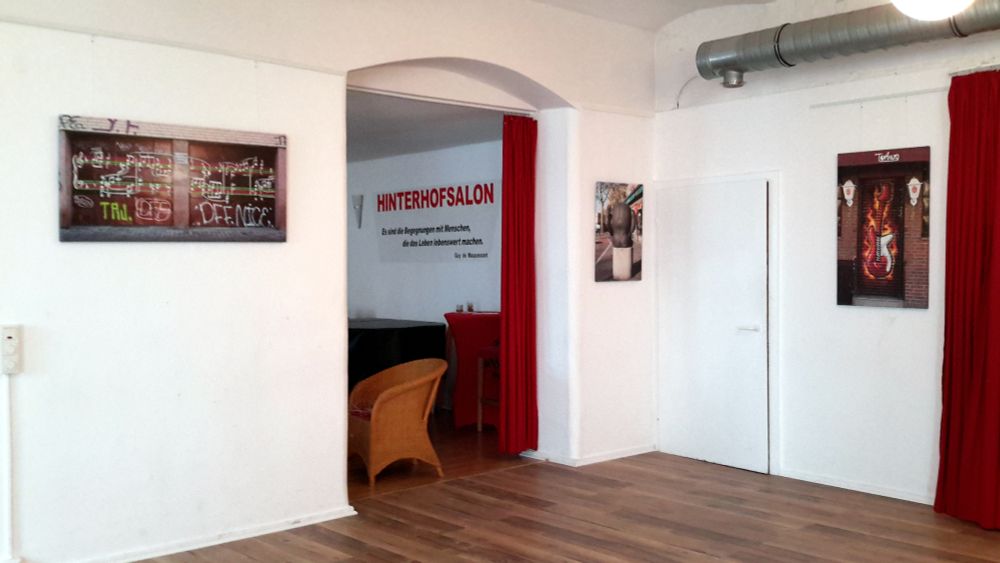 Raum mit Parkettboden und weiß gestrichenen Wänden, hinter dem rundbogigen Durchgang mit rotem Vorhang liest man die Schrift 'Hinterhofsalon'. Ein metallenes Rohr verläuft unter der Decke. An den freien Wänden hängen 3 Bilder: Eins mit einem Musiknoten-Graffiti, eins mit einer 'Arsch-mit-Ohren'-Skulptur, eins mit einer Tür und aufgesprühter E-Gitarre.