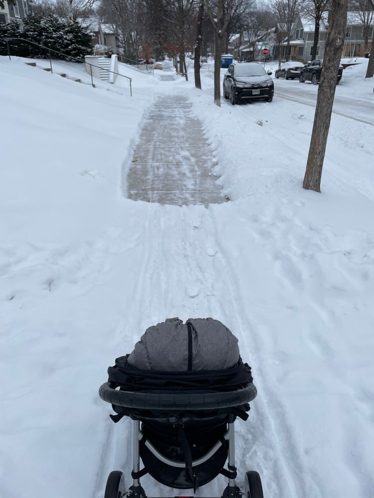 A stroller on a snowy sidewalk with some sections of the sidewalk clean in front of random houses. 