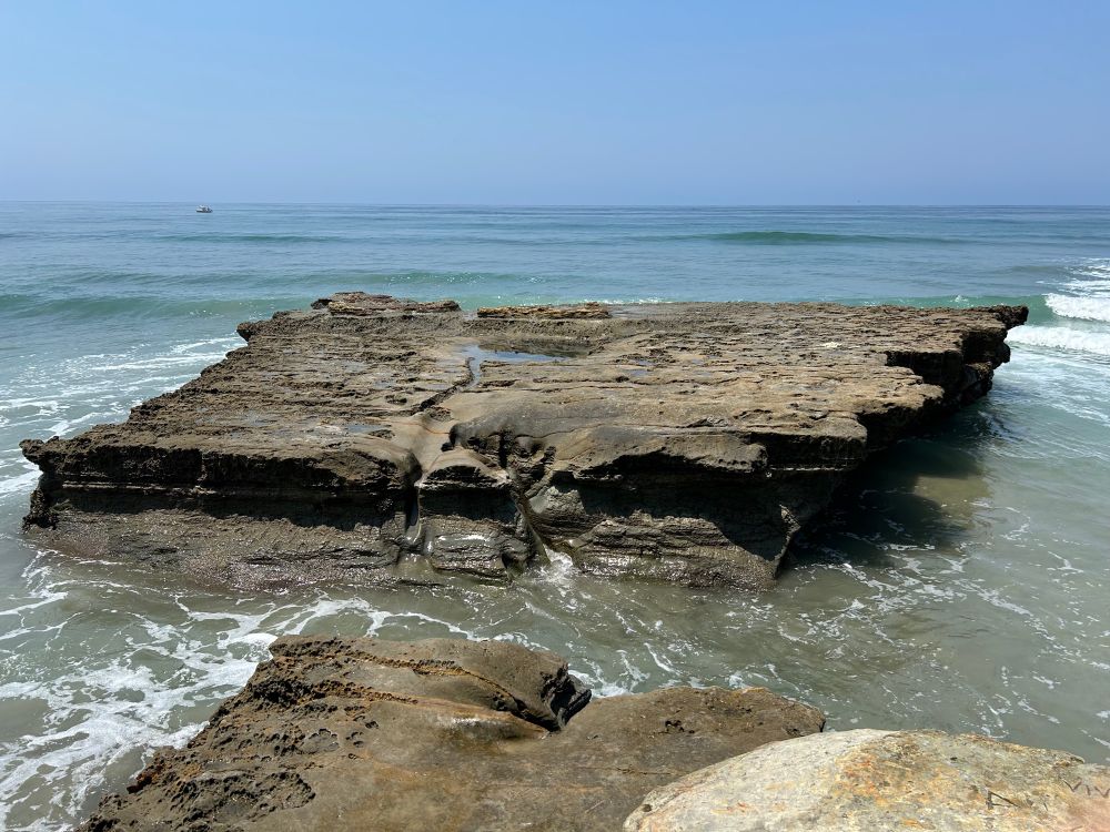 picture is of a large flat rock in a rough parallelogram shape, flat on top and about 6-8 feet above beach level. its surface is rough and pitted and if u you look closely there is a small square cavity in the center full of seawater. waves are rolling in, the ocean is turquoise close to shore, darkening to blue further out. the sun is overhead so there’s little shadow and no glare. lovely blue sky overhead 

this is Flat Rock at Torrey Pines Natural Reserve in San Diego, which separates Torrey Pines from the more notorious Blacks Beach to the south. 