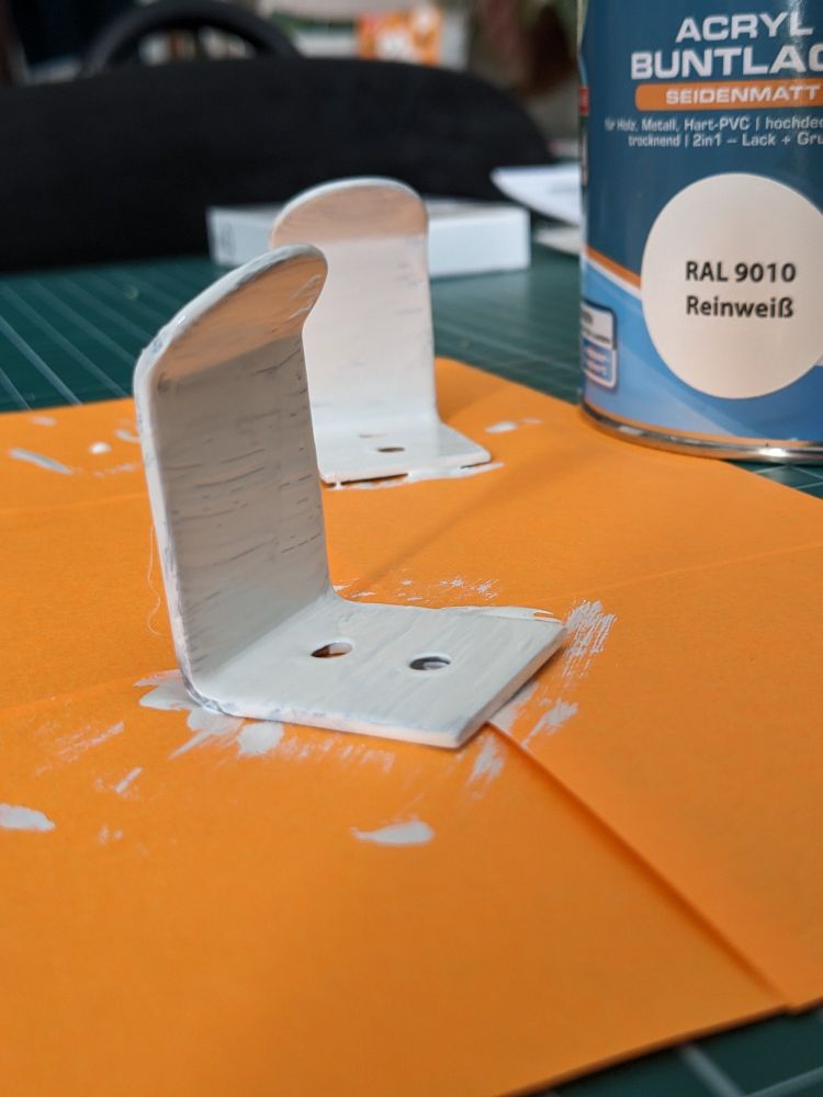 Nahaufnahme von frischlackierten  Wandhaken auf einer Unterlage, im Hintergrund eine Dose Weißlack