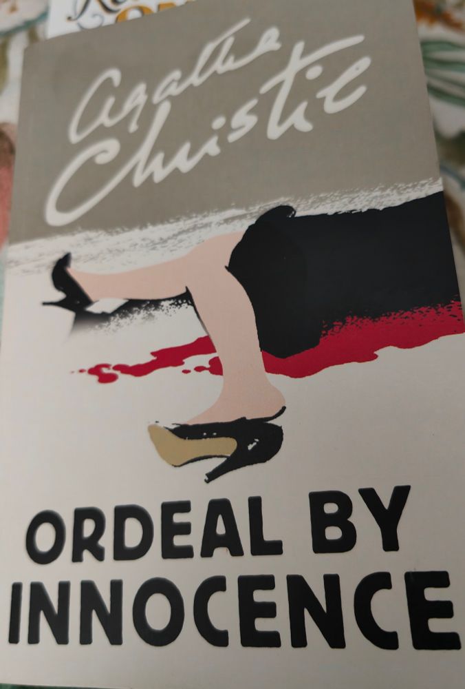 Front cover of Agatha Christie novel, Ordeal of Innocence. The cover image shows the lower half of a female wearing a black skirt and Stilleto black shoes, lying in a pool of red blood. 