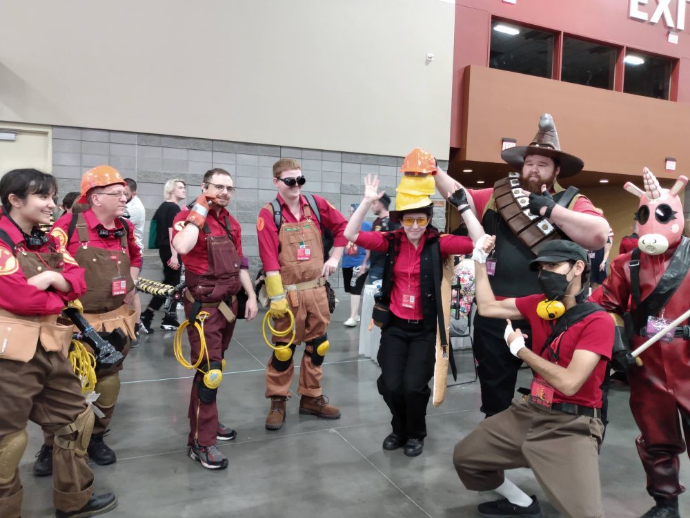 A crowd of cosplayers surrounding a sniper cosplayer with a tower of hats.