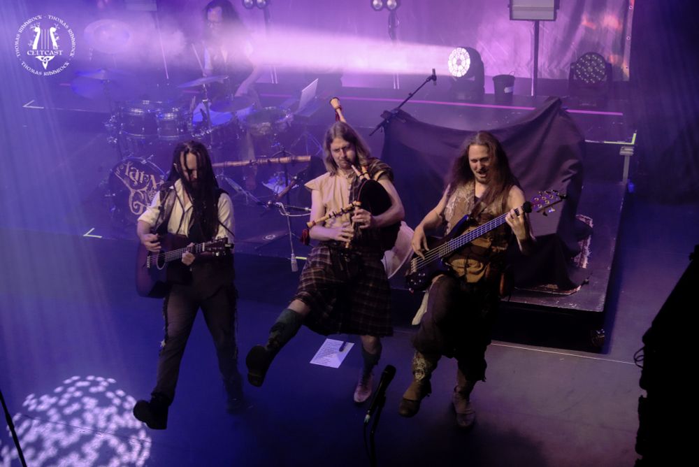 Image of Thomas (with guitar), Gilian (with bagpipes) and Dominique (on bass) dancing in a line at the front of the stage, all with the right leg up. Shot from the balcony right.