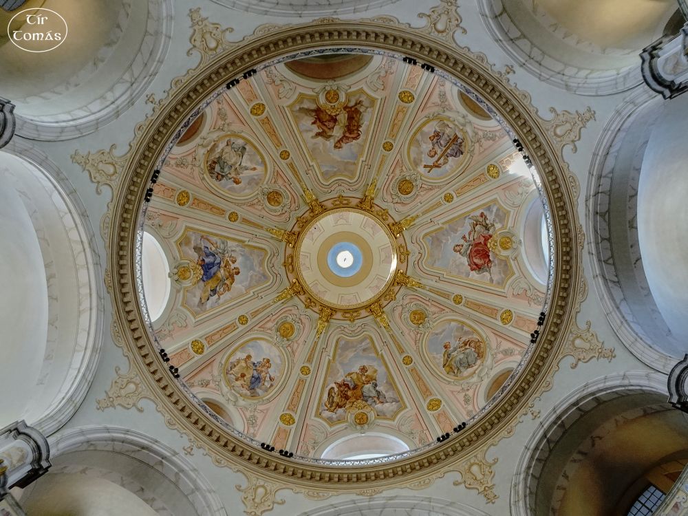 Round church dome from the inside - white surroundings, with a golden border at the edge of the dome and golden lines forming eight segments in the dome. Each segment has a biblical painting. In the very centre another golden ring, in the middle of which a small white spot of daylight can be seen.