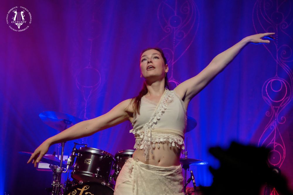 Image of Sophie van Grinsven, dressed in white, in dance, looking up, left arm pointing to the side up, right arm pointing to the side down. Drums in the backgound, and some decoration with blue and purple light can be seen.