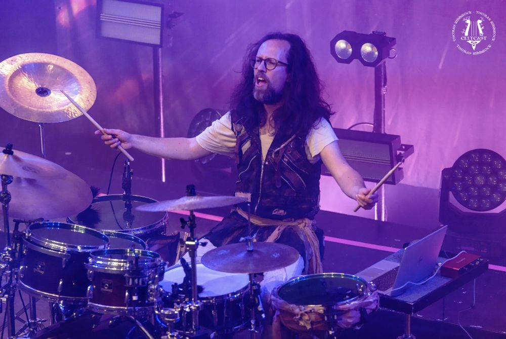 Image of Paul behind his drums, from the balcony, right looking down, arms wide with sticks in hands. He looks like he is shouting. Purple illumination.