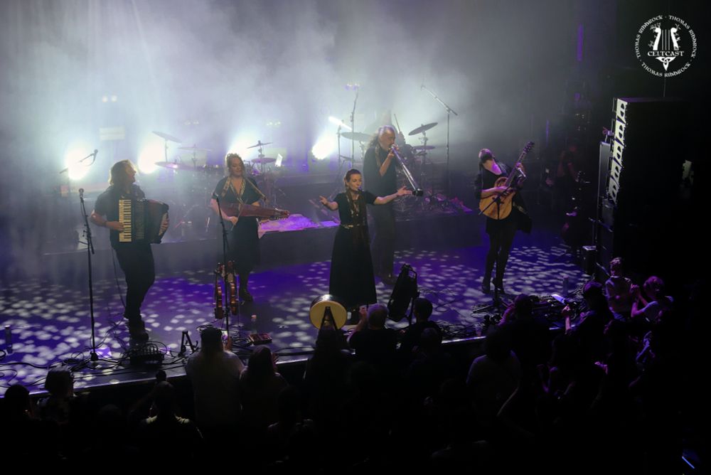 Image of the Bibelot-stage from the balcony - Cesair can be seen from a top-left angle. Left to right Daan on accordeon, Sohpie on violin, Monique singing with arms wide, Luka on slideridoo and Thomas on guitar. In the background on the right, the drumset is barely visible in the smoke. In front of the stage, the silhouettes of people can be seen.