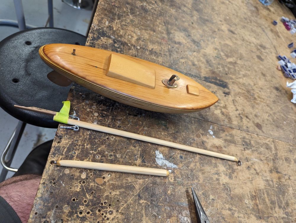 A small wooden toy sailing boat on a dirty bench with a wooden mast and boom laying next to it. The bench has a bunch of drill holes in it that I wish weren't the inevitable result of a shared makerspace workshop. Use a bit of scrap wood people!