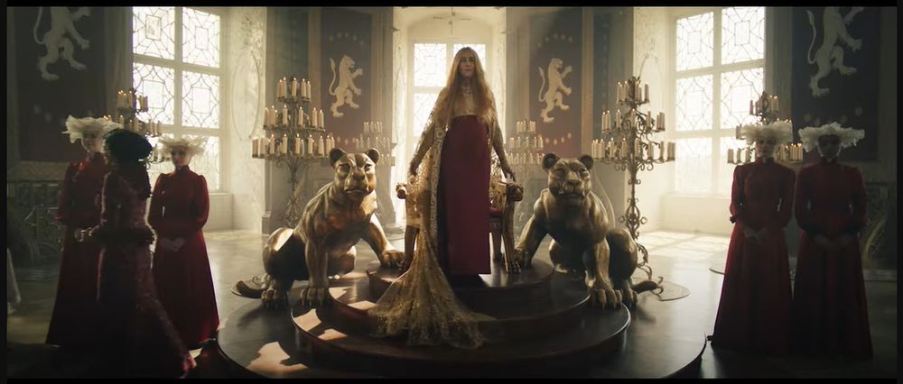 A closer shot of the Lion Throne with Queen Morgase standing before it at the top of the steps leading to it. She is wearing a red dress with a flowing golden shawl. 
The Throne itself is a large wooden chair with life size golden lionesses on either side.
There are White Lion banners and large windows behind the throne. 
There are two maids in red dresses and white head dresses each side of the throne (No not like the Handmaids Tale).
