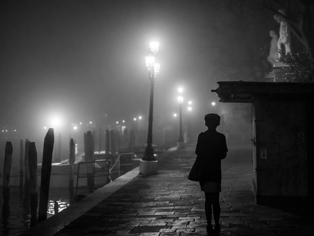 A black and white image of a lone woman walking in the fog in Venice, with her back to us, illuminated by faint street lights. 