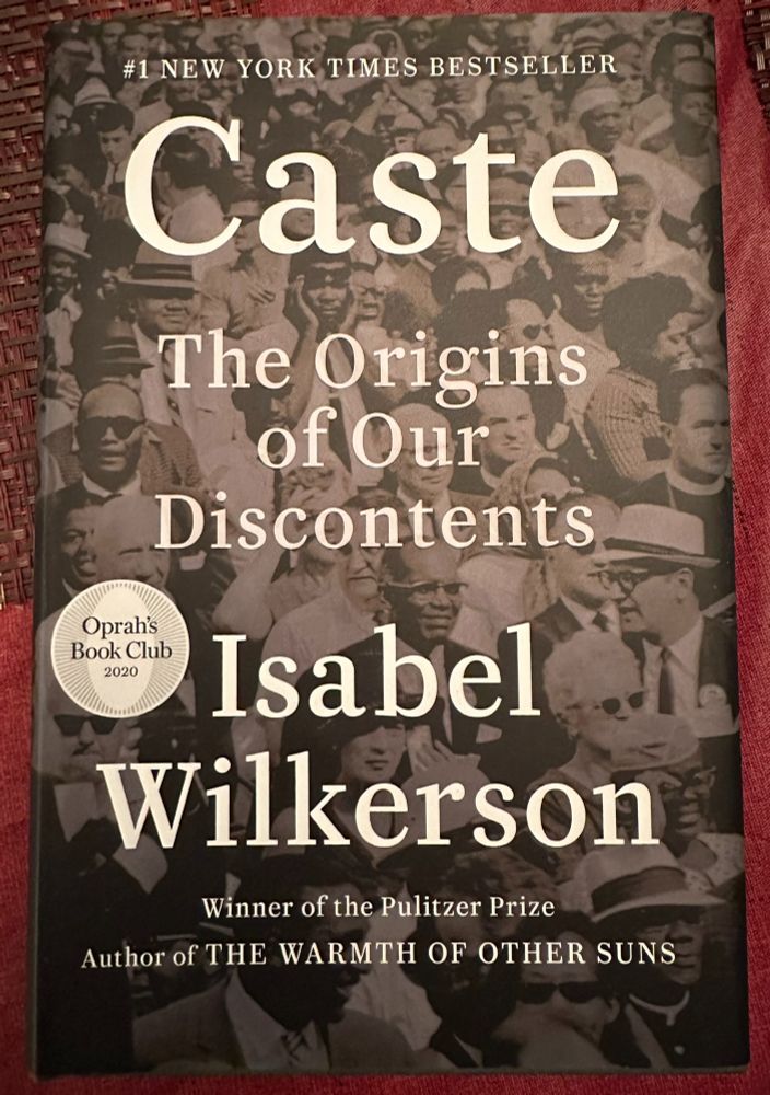 Image of the front cover of Isabel Wilkerson's book, 'Caste: The Origins of Our Discontents.' The cover features a black and white photo of a diverse crowd and the title/author in large white text. It also notes Wilkerson is a Pulitzer Prize-winning author and this book was an Oprah's Book Club selection (2020). Wilkerson is also the author of 'The Warmth of Other Suns.