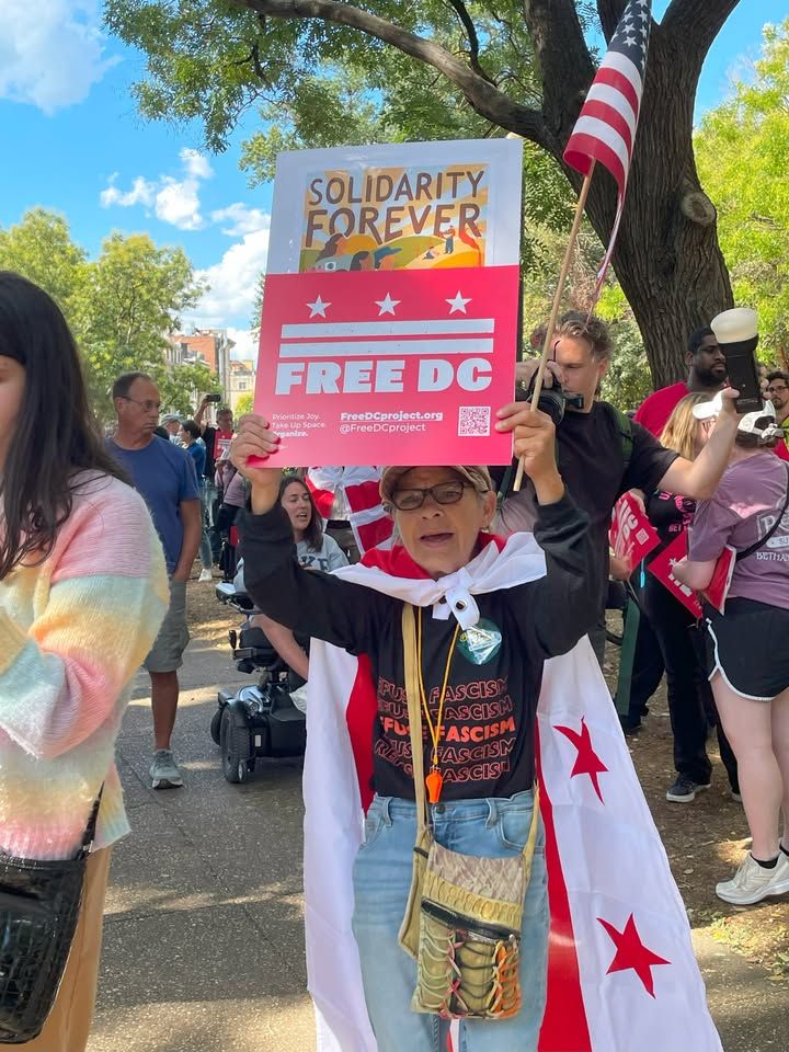 A person holding a 'Solidarity Forever FREE DC' sign at a public demonstration, draped in a flag with white stars on a red background.