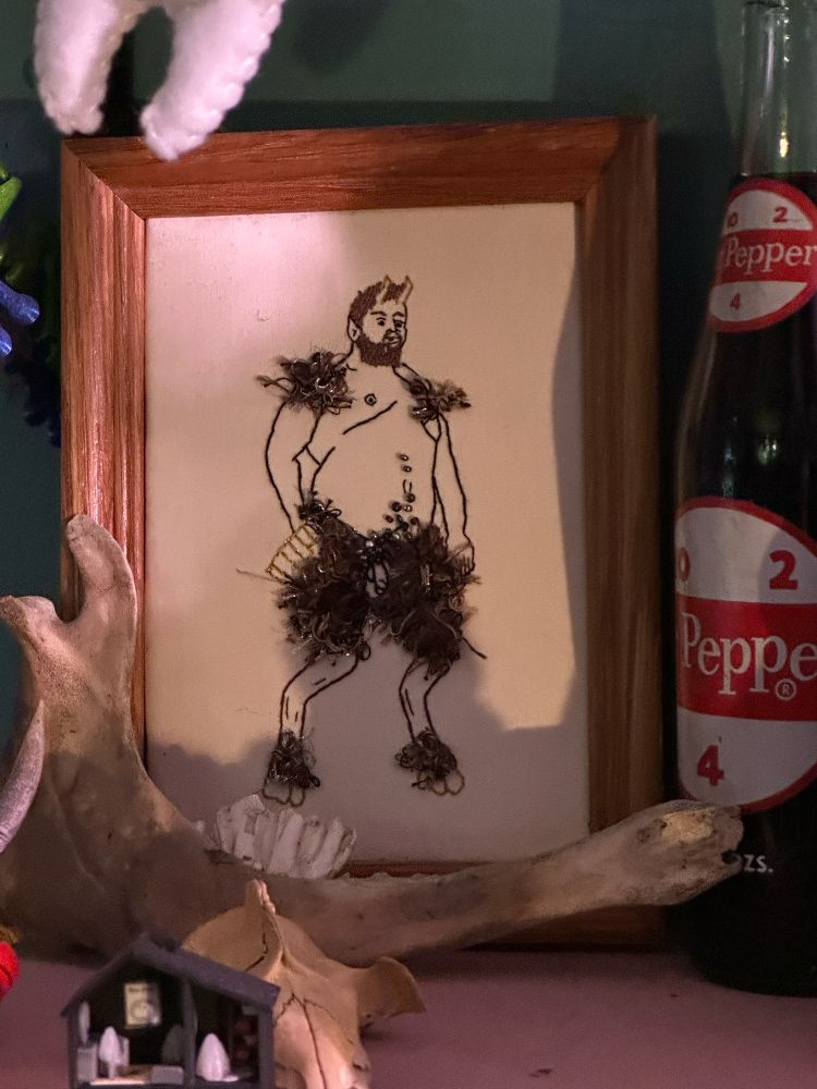 An embroidery of a fat male presenting person turned into a satyr. Various shades of brown and silver threads are used to represent the fur 
