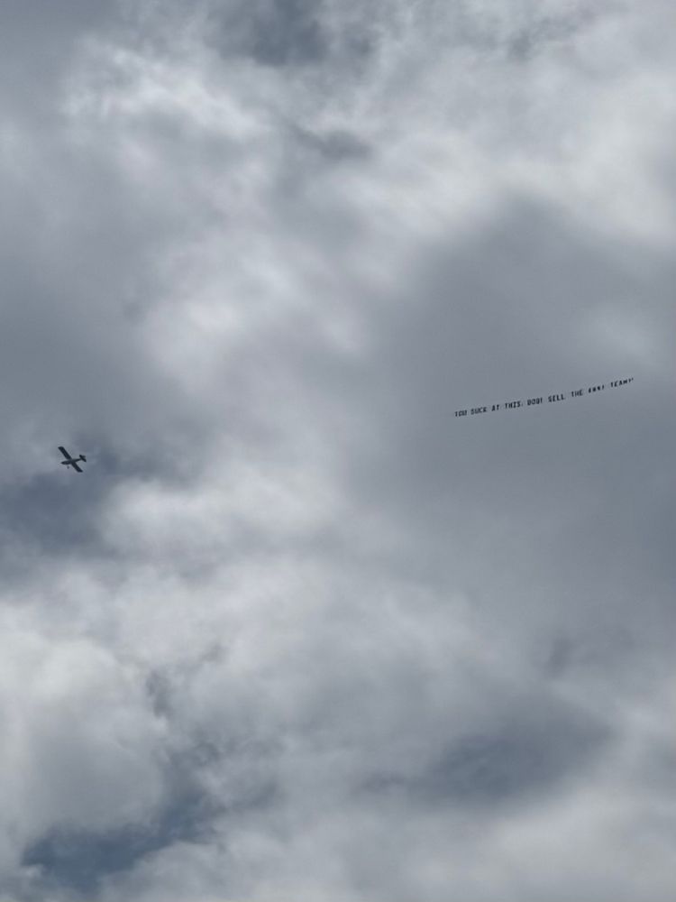 Picture of a mostly cloudy sky with a plane silhouetted against it, flying a banner behind it