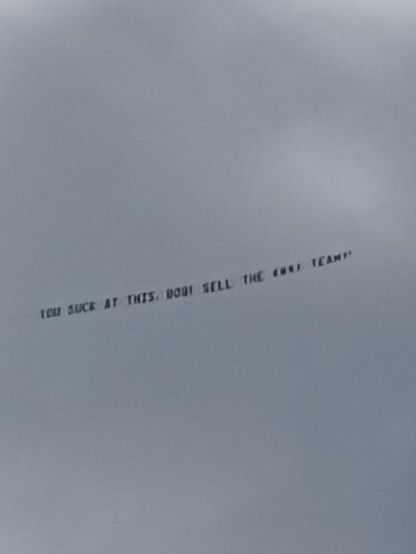 Plane banner against clouds that reads “YOU SUCK AT THIS, BOB! SELL THE @%*! TEAM!!”