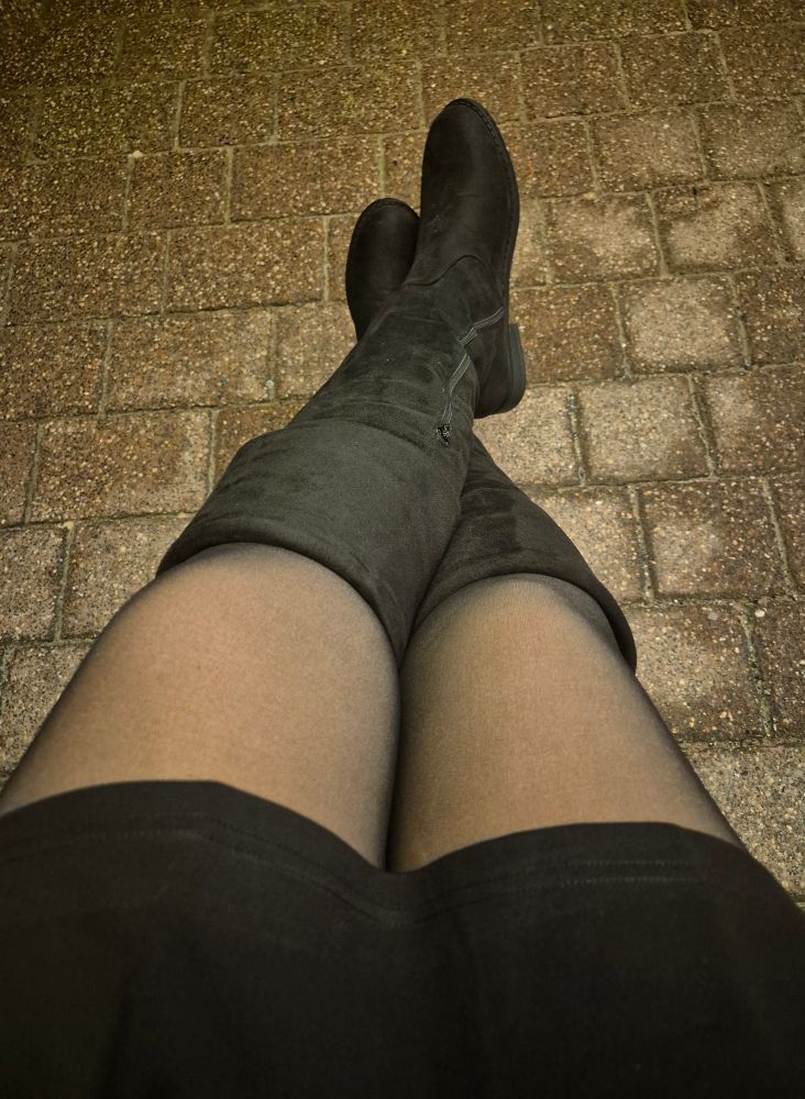 Die Beine der Userin in einem kurzen schwarzen Kleid, schwarzen Strumpfhosen und schwarzen kniehohen Striefeln.