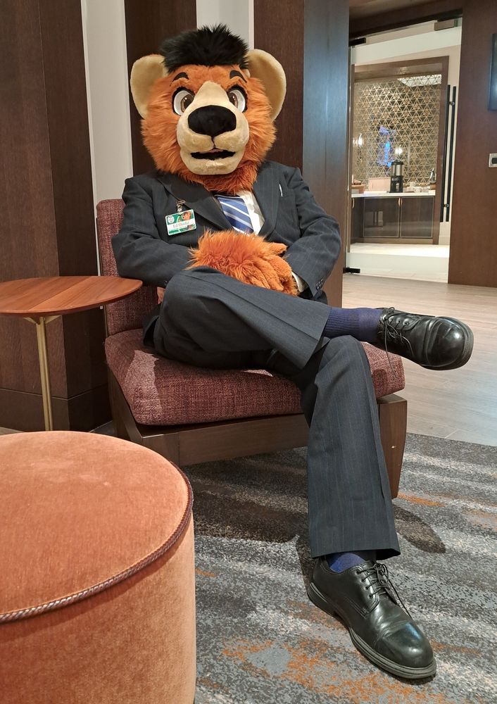 A man in a business suit sitting in a chair in a hotel lobby.  He is wearing a bear fursuit head and handpaws.
