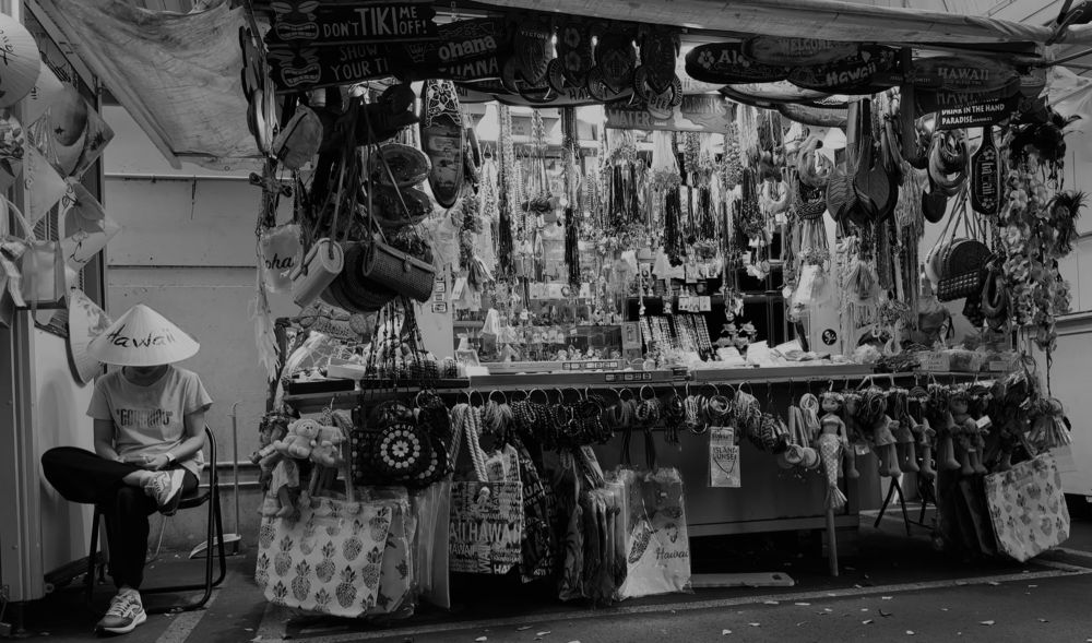 Black and white photo of an open air vendor stall full of tacky Hawaiiana - shell necklaces, novelty tikis, etc. The proprietor sits nearby in a folding chair, on their phone, wearing one of the products - a pointed straw hat with "Hawaii" printed on the front in script. 