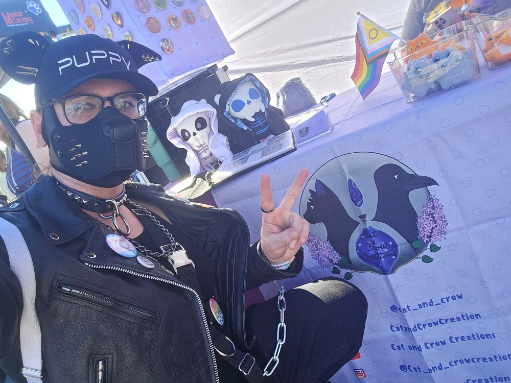 Me in my black spiked pup gear kneeling by my fursuit maker's booth at Pride in the Park