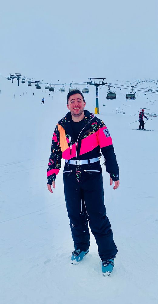 Me standing at the bottom of a ski slope, smiling and wearing a black ski suit with pink and orange fluorescent patterning on the top half.