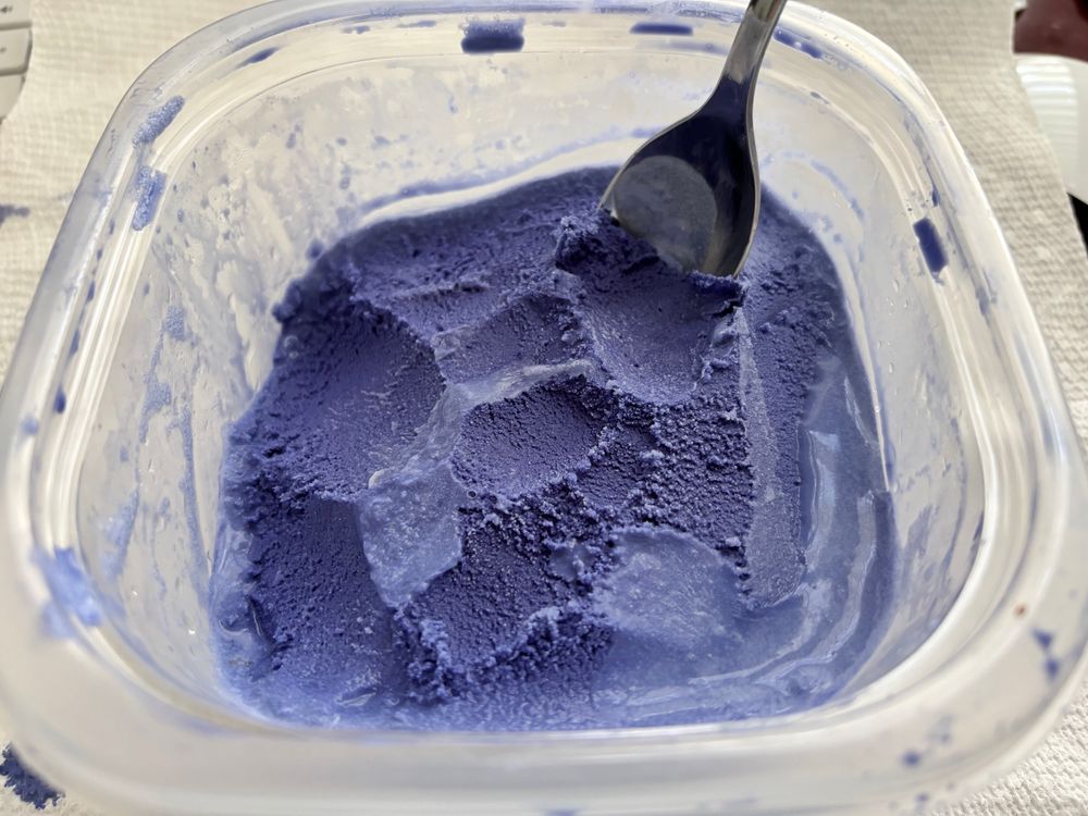A clear plastic container about 2/3-3/4 filled with bright purple ice cream. A spoon has been stuck into the upper right corner.