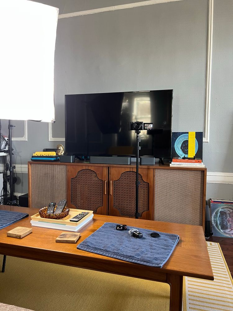 Photo of my livingroom setup. In the frame is a large photography light, a camera on a tripod and microphone equipment sitting on a coffee table. 