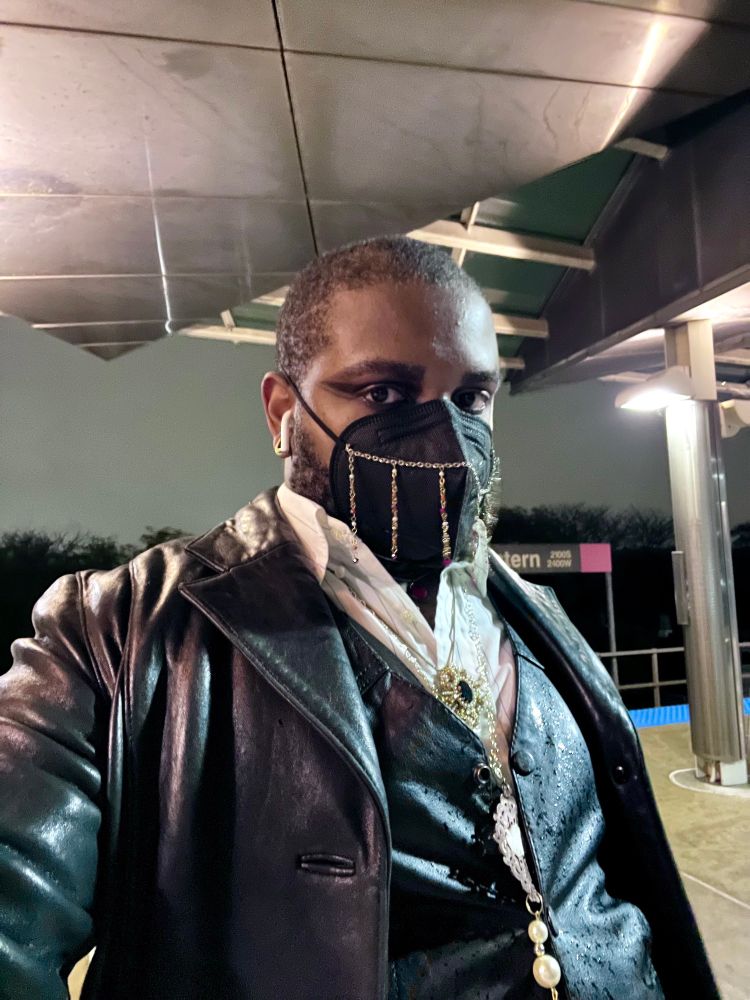 Selfie of me on a Chicago train platform. I’m dressed in a leather vampire-esque outfit and wearing a black N95 respirator adorned with jewelry. 