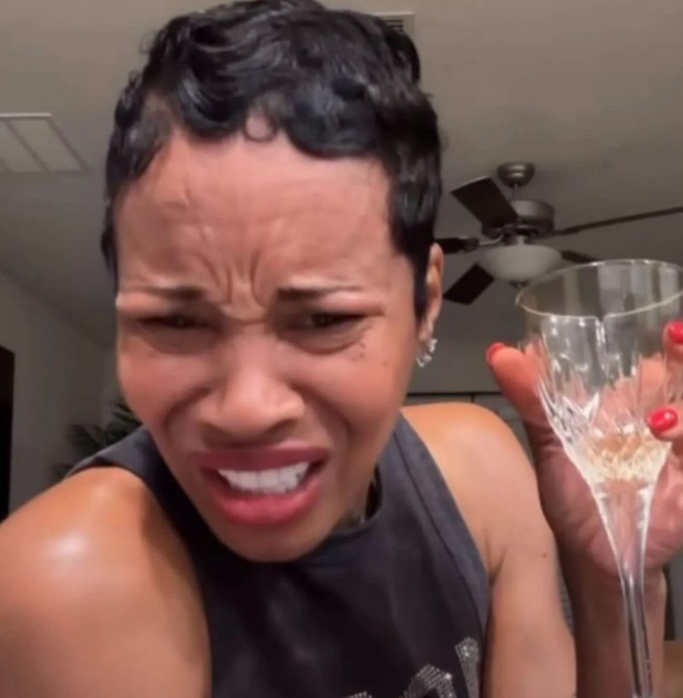 Photo of a black woman with a disgusted confused expression, holding a crystal glass. She has short furled hair and is wearing a black tank top