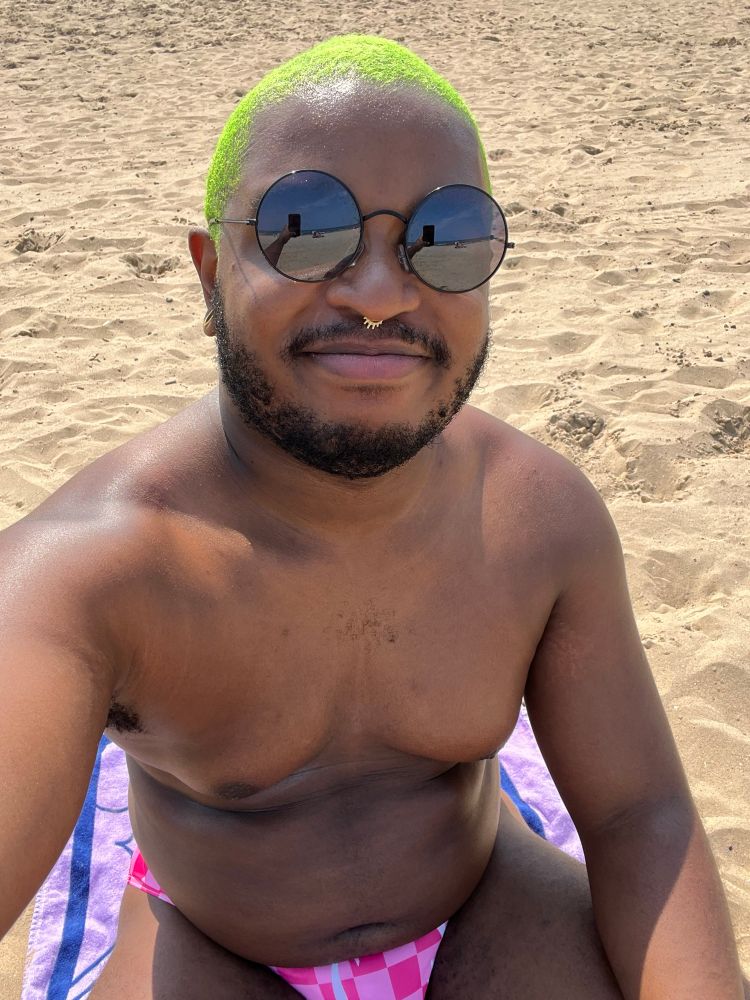 Selfie of myself sitting at the beach. I’m wearing a pink swimsuit and circular sunglasses 
