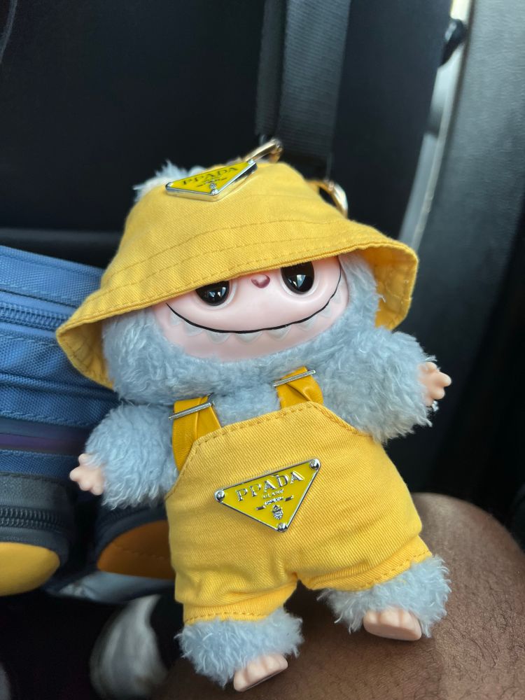 A light blue labubu plushie wearing a yellow Prada bucket hat and a matching pair of overalls. 