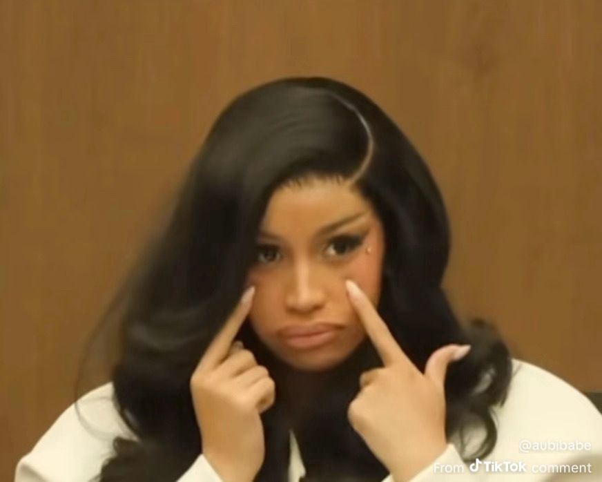 Photo of Cardi B sitting on the stand in a courtroom. She’s pointing to her eyes and making a puppy dog eyes sorta face, with a tinge of confusion. 