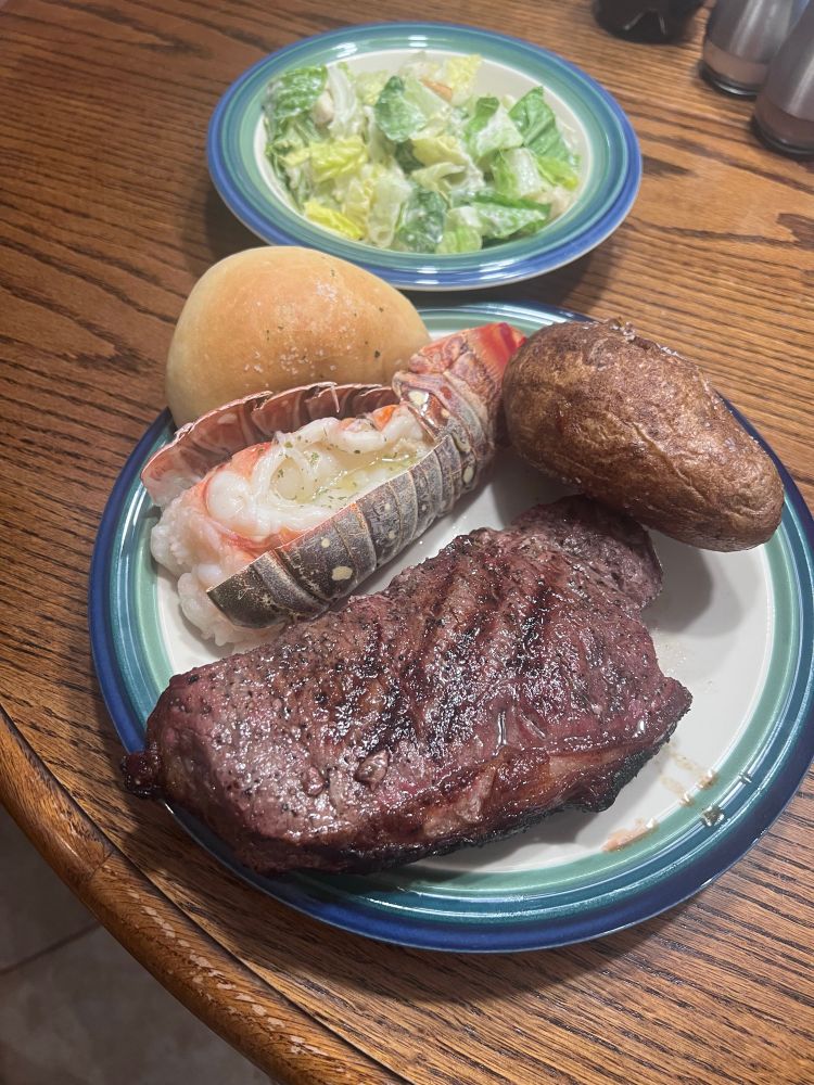 A picture of a New York strip steak, baked potato, lobster tail, roll, and Caesar salad.
