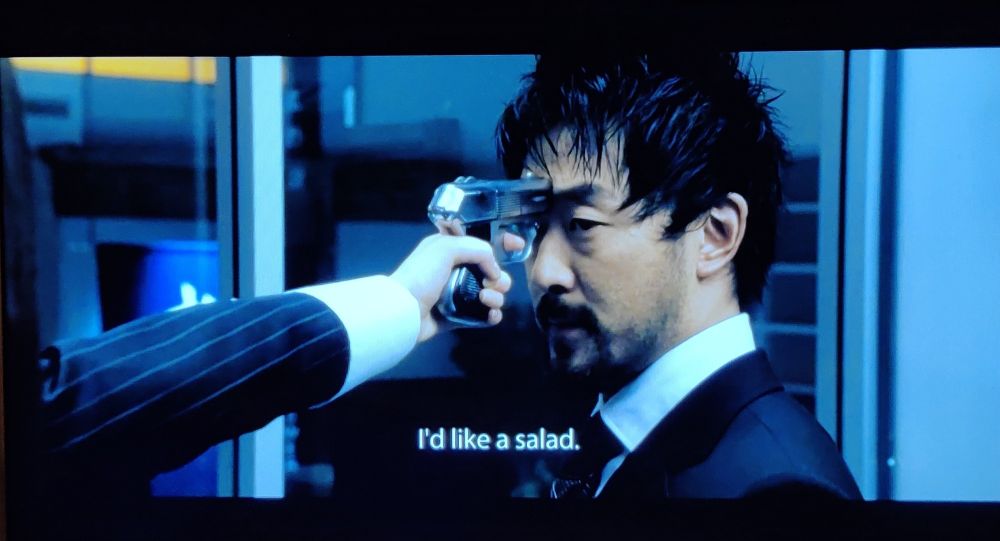 A woman's hand hold a gun to a man's head. They are both in suits. The subtitles show the woman is saying (offscreen) "I'd like a salad."