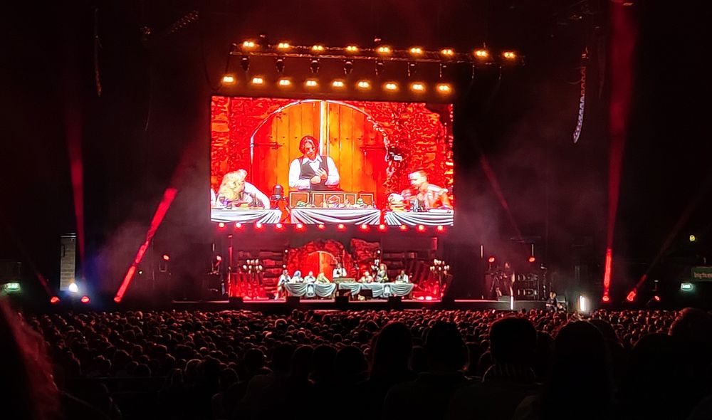 A view of the stage during the Critical Role show. The stage and cast are visible, beneath a huge screen showing Matthew Mercer standing behind his GM screen. A sea of heads is visible below, barely highlighted by the stage lights. Probably a couple thousand people visible even in this limited view.