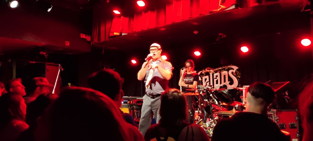 MC Frontalot in classic stage uniform of office wear, white tie and forehead torch like cyclists wear? Performing on stage at Whelan's, Dublin. Accompanied by Lauren Ipsum, in much more casual t-shirt and jeans. Red lights, audience in the foreground.