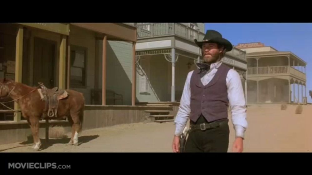 a man in a cowboy hat is standing in front of a building that says ' saddle maker ' on it