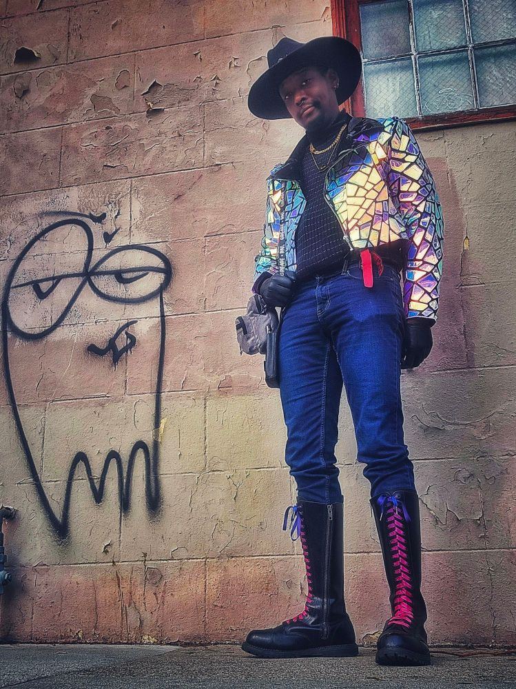 A man in tight blue jeans, tall boots, a shiny jacket and a cowboy hat stands against a city wall with graffiti that looks like a ghost with half-opened eyes.