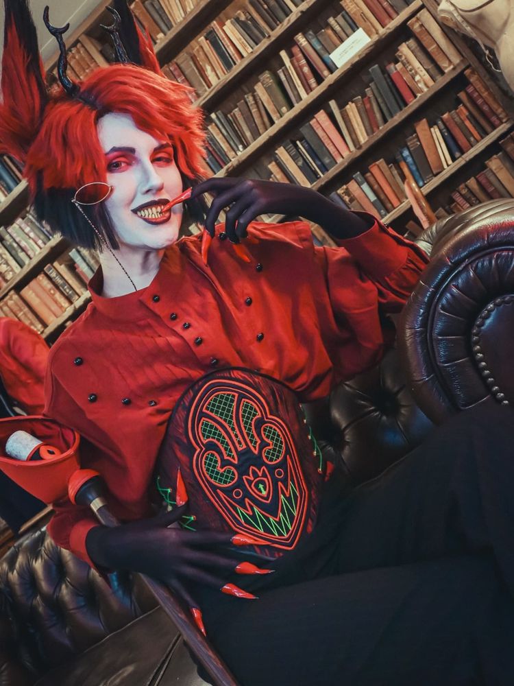 Chari dressed as Alastor wearing a loose fitting red shirt and black and red waist cincher with a red and neon green radio designed and embroidered by Arty. He's sitting on a brown leather sofa on front of a large bookcase. He's holding his microphone cane loosely in one arm while smiling and biting the tip of his nail on the opposite finger.  