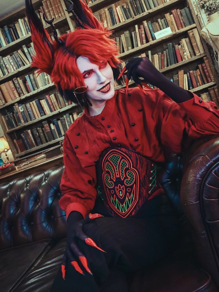 Chari dressed as Alastor wearing a loose fitting red shirt and black and red waist cincher with a red and neon green radio designed and embroidered by Arty. He is looking thoughtfully to the side at something off camera. End ID)