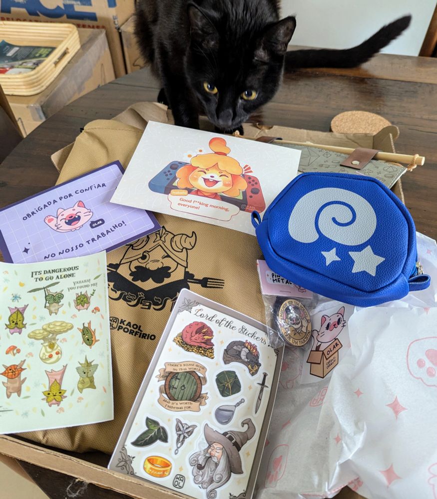 A black cat sniffing at anime goods from an independent retailer's shop. Items include stickers, pins and an apron from various games, movies, and anime, including Lord of the Rings, Porco Russo, Animal Crossing, Delicious in Dungeon, and Zelda.