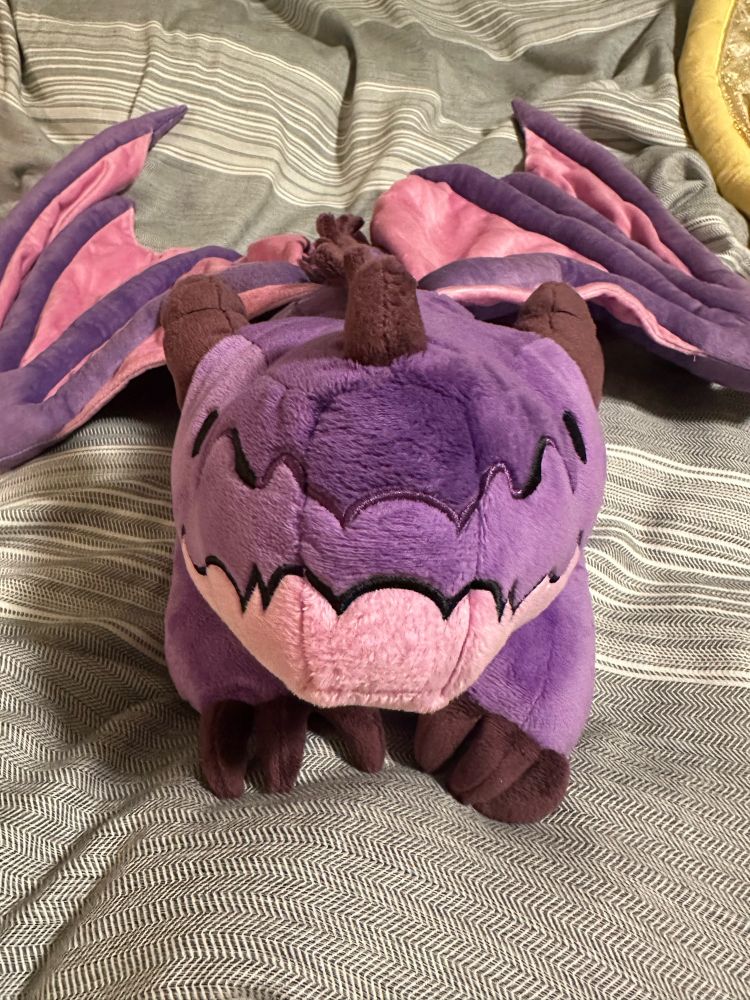A purple warm-able dragon plush looking directly at you from Weighted Wonders.