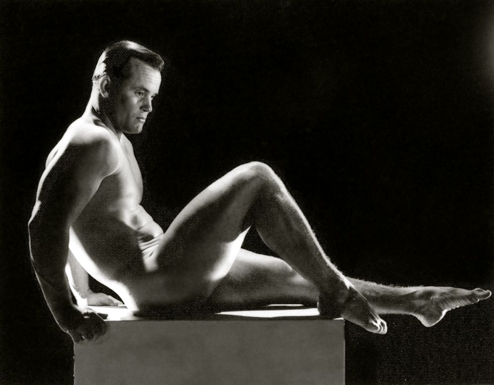 Black and white photo of the outline of a backlit nude male against a black backdrop with the lights directly behind him.